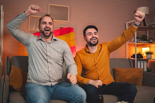 Two friends are joyfully cheering while seated on their sofa, surrounded by a cozy home atmosphere. A vibrant flag decorates the backdrop as they enjoy the excitement of the game.