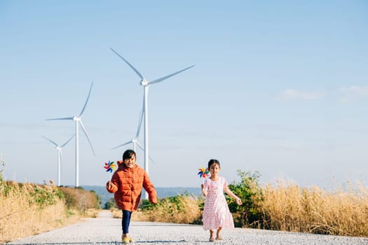 A girl and her sister holding pinwheels run by windmills. Family bonding near turbines signifies clean energy. Children joy windmill circle against clear sky reflects sustainable cheerful future.
