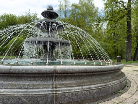 Close up of stone fountain with layered tiers and arched water jets in public park. Clear focus on textured surface and dynamic water flow design