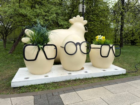 Decorative sculpture of hen and three egg shaped flower pots with black glasses on wooden platform in city park. Whimsical urban art installation with humorous design