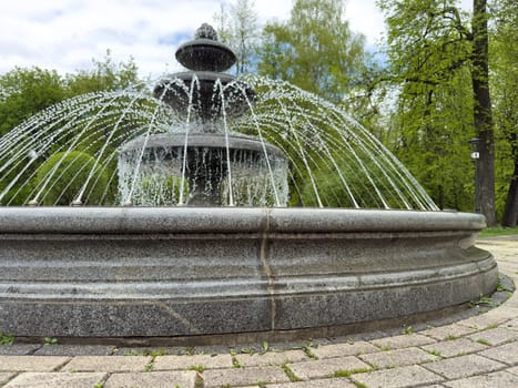 Classic round granite fountain with decorative multi-tiered structure and dynamic water jets in urban park environment. Public landscaping concept with stone tiles and green background