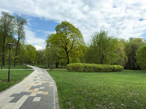 Beautiful city park with modern patterned walking path, lush green trees and bushes, and streetlights along the trail. Peaceful natural environment perfect for recreation and relaxation