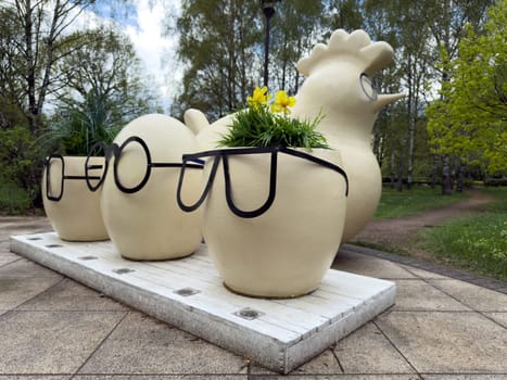 Creative public art installation featuring planters in shape of chicken eggs wearing eyeglasses next to a stylized chicken sculpture. Unique urban landscaping idea for outdoor visual attraction