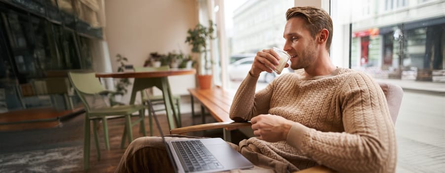 Portrait of handsome modern man, sitting in coffee shop with laptop, looking at screen with happy face, drinking cappuccino, resting in cafe or working.