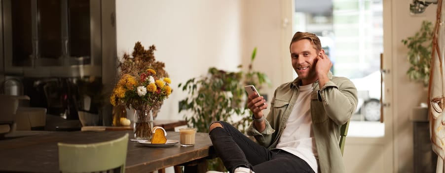 Happy handsome man in coffee shop, listening to music in wireless earphones, connects to cafe wifi to enjoy favourite song on his smartphone playlist, sitting and drinking cappuccino.
