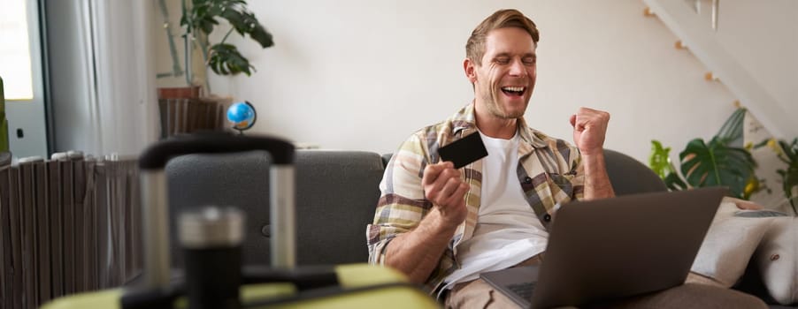Portrait of happy man, traveller, holding credit card and using laptop, booking vacation, buying tickets online for summer holiday, has suitcase packed.