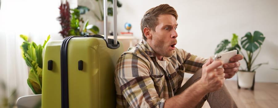 Man with suitcase, sits on floor, looks shocked at his phone screen. Tourism concept