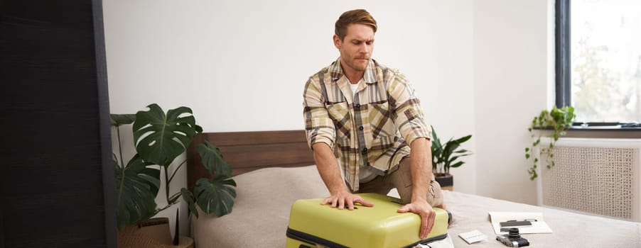 Portrait of young man, tourist packs his bag, closing full suitcase, trying to push the luggage inside to zip the bag, sitting on bed in bedroom. Tourism and adventure concept