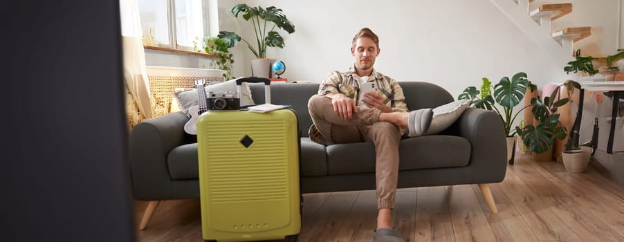 Portrait of man sits with mobile phone in living room, packed suitcase, chats on smartphone app, looks for holiday destinations, goes on vacation.
