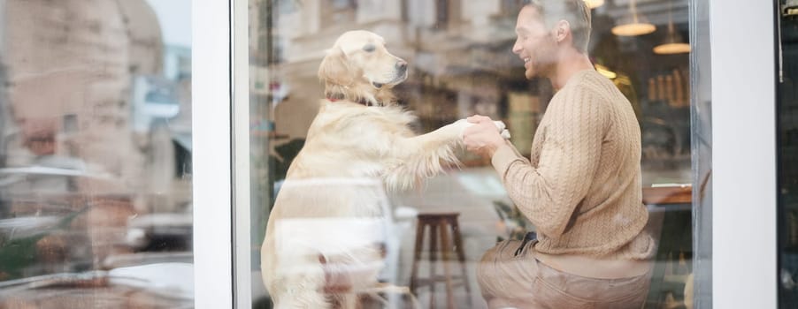 An outdoor shot of a man sitting with his dog in a pet-friendly cafe near window, golden retriever gives paw to visitor.