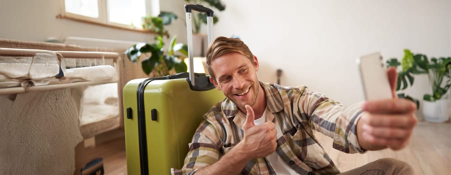 Happy young man shows thumbs up, takes selfie on mobile phone, poses near suitcase, blogger goes on holiday and posts about vacation. Tourism concept