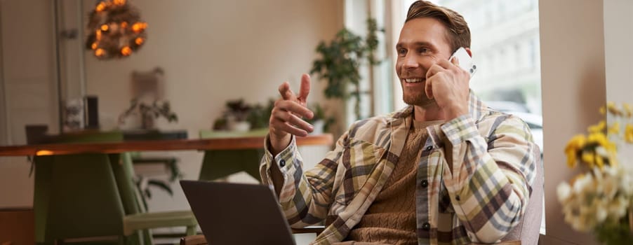 Handsome businessman in coffee shop, sitting in cafe with laptop and calling someone, Man talking on the phone with client, making enquiry about project, talking to coworker while working remotely.