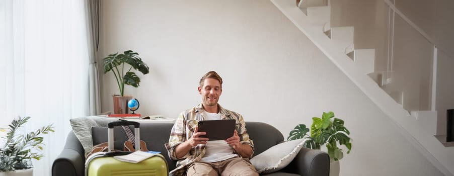 Vertical shot of man, tourist with suitcase, looking at digital tablet, reading about travel destination, booking hotel on gadget. Tourism concept