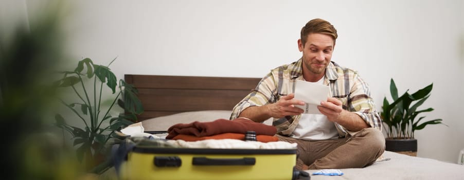 Portrait of excited young man, tourist going on vacation, has his tickets, sitting near opened suitcase, looking at passport with pleased smile. Lifestyle concept