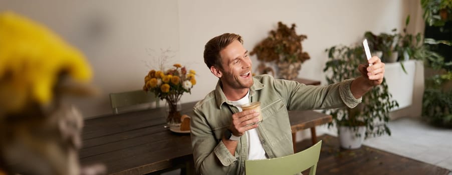 Portrait of handsome happy young man, taking selfie in cafe, posing with cup of coffee, video chats using smartphone app, talks to a friend. Lifestyle and leisure concept