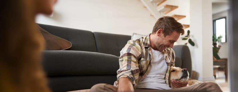 Happy young smiling guy in living room, sitting on floor, hugging his pet, playing with dog. People and animals concept