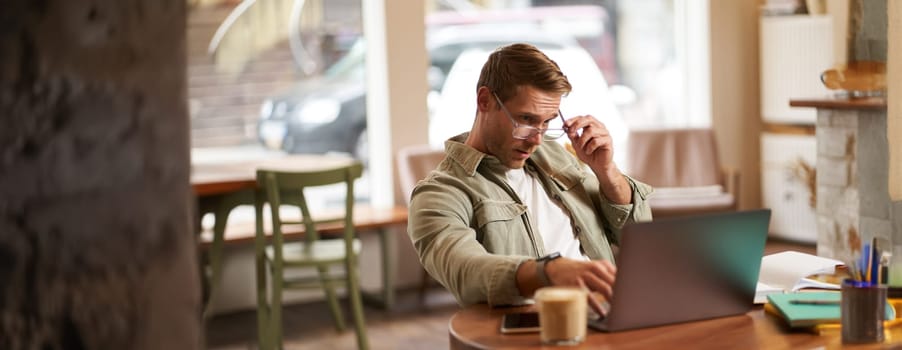 Portrait of a man working in cafe, looking surprised at camera, taking off glasses to stare puzzled at laptop screen, has a question about something on his computer. concept of remote jobs.