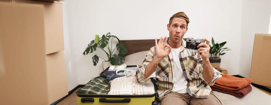 Portrait of young handsome man, travel photographer, shows okay sign, sits with camera on bed, packing things in suitcase, going on holiday or vacation abroad.