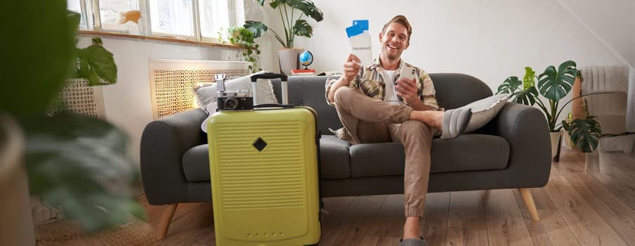 Portrait of young tourist, man on sofa with suitcase, preparing for holiday, sitting with smartphone, booking hotel and plane tickets online on mobile phone app. Tourism and travelling concept