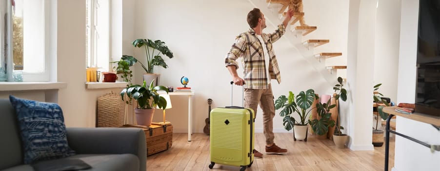 Portrait of happy young tourist, man walks indoors with suitcase, pets his cat. Tourism and travelling people concept