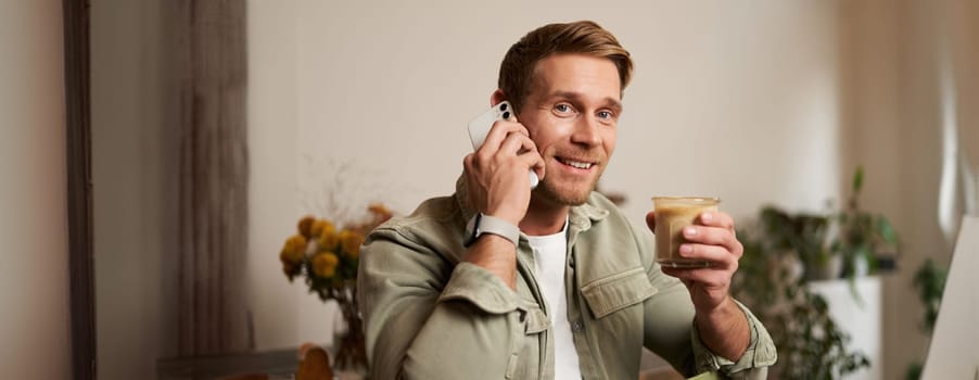 Image of smiling, good-looking young blond man with phone, drinking coffee in cafe, talking to someone over the telephone, answering a call.