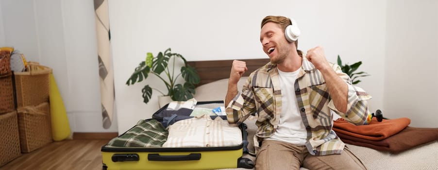 Portrait of cheerful handsome man, tourist going on vacation, dancing and listening to music in headphones while packing suitcase with clothes for holiday trip abroad.