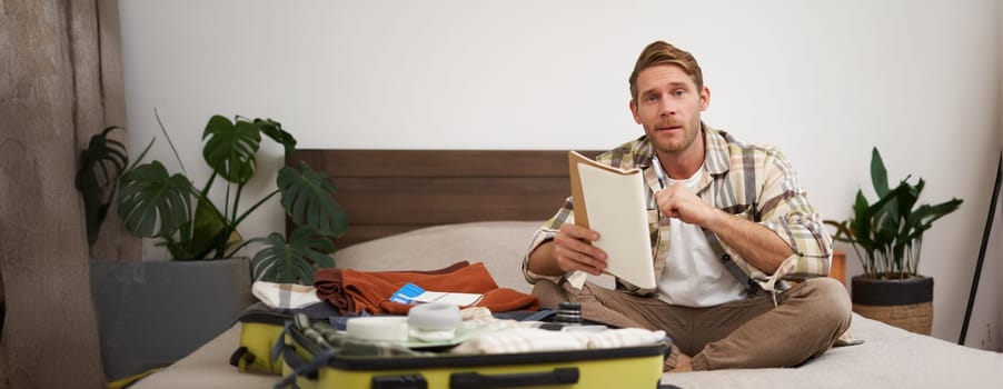 Portrait of young man, looking happy, going on holiday trip, travelling, holding notebooks with list of items, packing suitcase while sitting on bed.