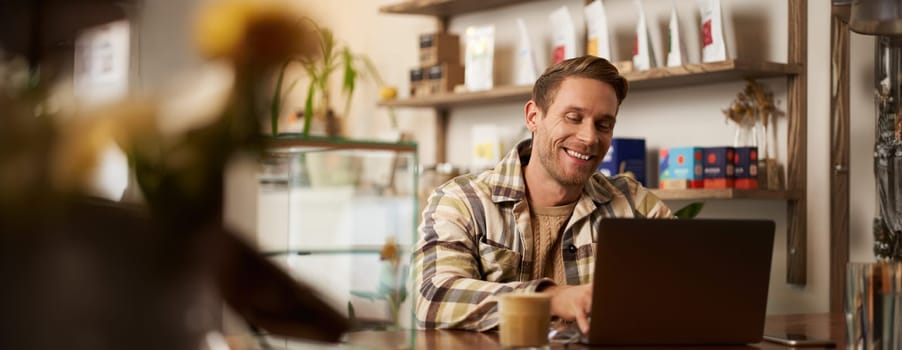 Portrait of handsome young man using laptop in cafe, sitting with computer in creative public space, typing on keyboard with pleased happy smile.