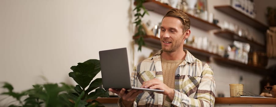 Portrait of young man working online, freelancer sitting in cafe, doing his project on laptop, drinking coffee, smiling while looking at screen.