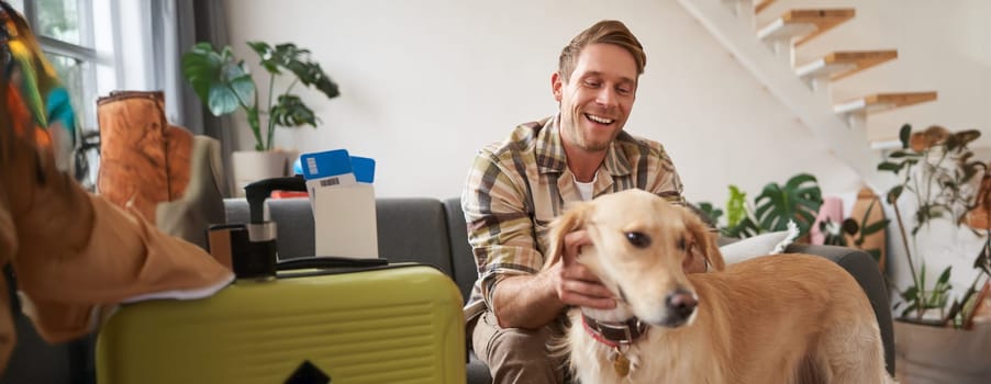 Portrait of happy, handsome young man with suitcase, sitting with his dog in living room, prepared to go on vacation, going on holiday. Tourism concept
