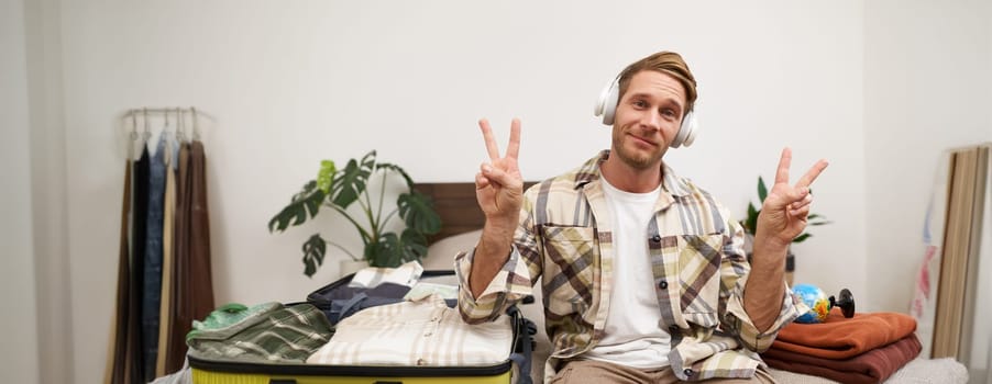 Portrait of cheerful handsome man, tourist going on vacation, dancing and listening to music in headphones while packing suitcase with clothes for holiday trip abroad.