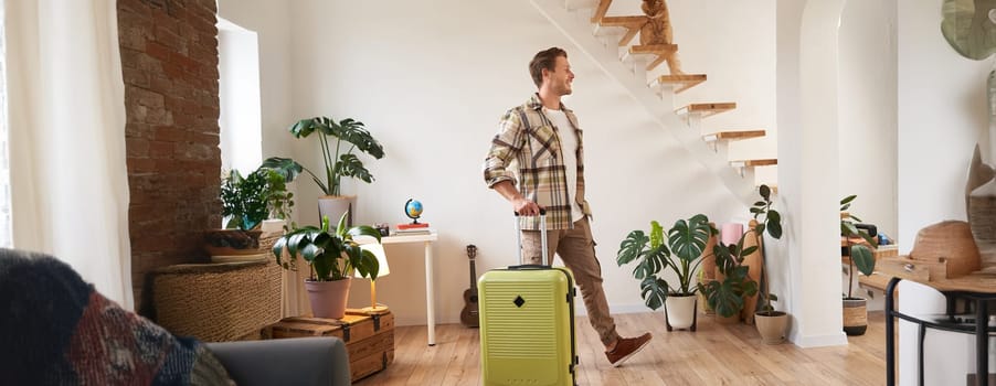 Portrait of handsome young man walking with suitcase in house, renting a room on vacation, smiling and looking happy. Travelling and tourism concept