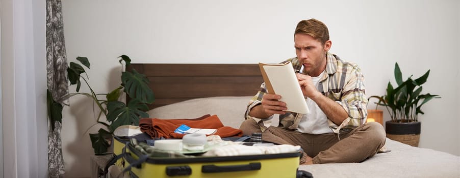 Portrait of angry man, packing suitcase, looking frustrated at notebook with check list of items for holiday, preparing to go on vacation. Tourist and people concept