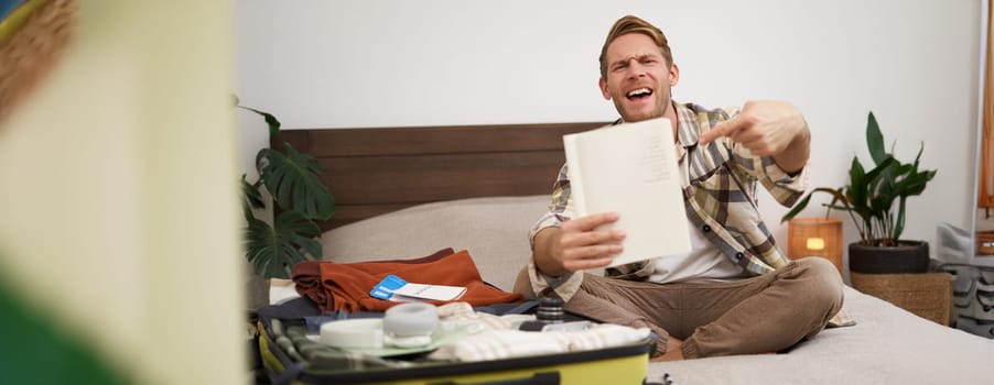 Travelling and tourism. Young man sits on bed, shows his list of items, demonstrates notebook, packs clothes, prepares to go on holiday, goes on vacation, assembles luggage.