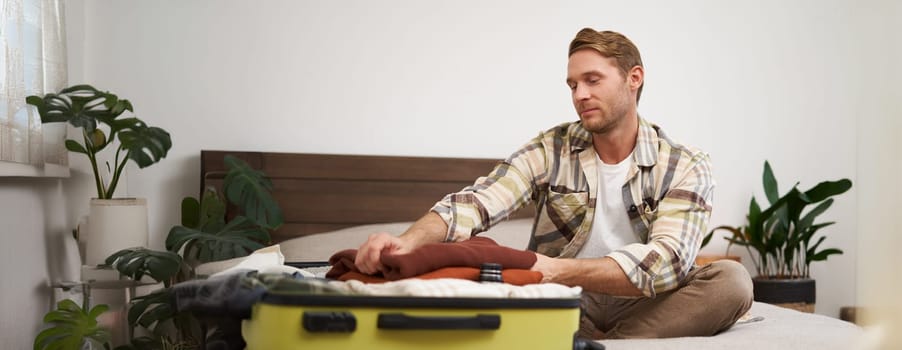 Portrait of young man packing clothes in suitcase, sitting on bed with luggage, going on vacation, preparing for a journey. Lifestyle and tourism concept