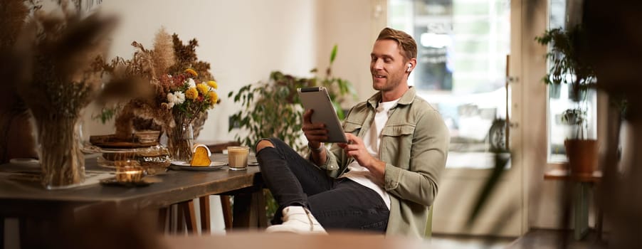 Portrait of young man sitting in cafe, spending time alone in coffee shop, reading on digital tablet, watching video, listening music in wireless headphones. Lifestyle and people concept.