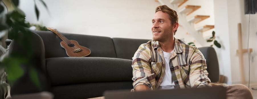 Close up portrait of young man using laptop, looking at computer, sitting on floor with his gadget, browsing websites.