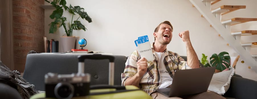 Enthusiastic young man, tourist looking excited, saying yes, holding two tickets, sitting with laptop and screaming from happiness, has suitcase and camera prepared for holiday.