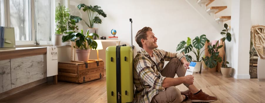 Portrait of smiling handsome tourist, man holding passport and flight tickets, sitting with suitcase. Tourism and travelling concept