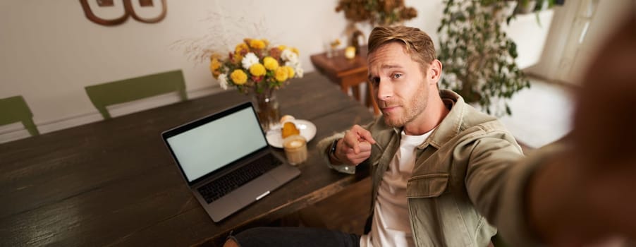 Handsome and confident young man taking selfie on mobile phone, pointing finger at camera, sitting in cafe with laptop, working from coffee shop, looking sassy.
