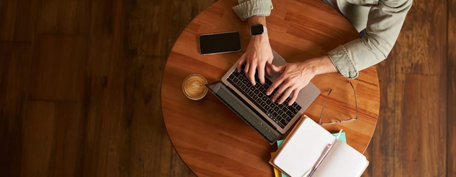 Cropped top view of male hands, man typing on laptop keyboard, working in cafe, sitting at round table in coffee shop, has phone, notebook and cup.