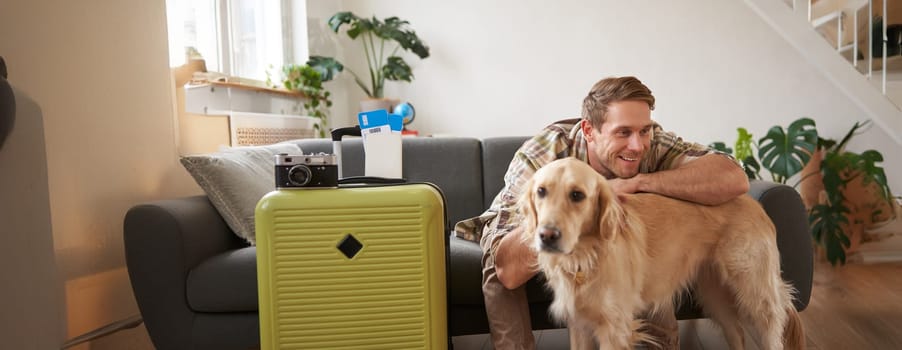 Portrait of happy young man going on holiday, travelling with his dog companion, sitting with golden retriever near suitcases and tickets.