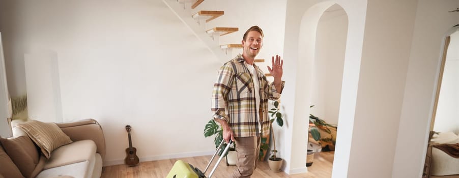 Full length photo of happy young man, tourist going on holiday, traveller leaving apartment with suitcase, waving hand at camera to say goodbye. Copy space