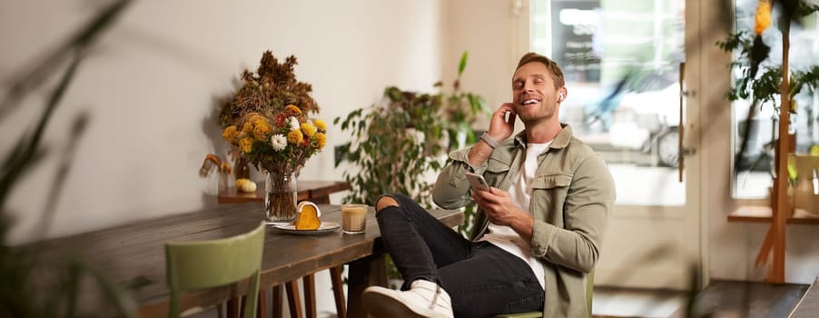 Portrait of handsome happy young man, relaxing in a coffee shop, spending time in cafe, listening to music in wireless headphones, holding smartphone, putting on his favourite song.