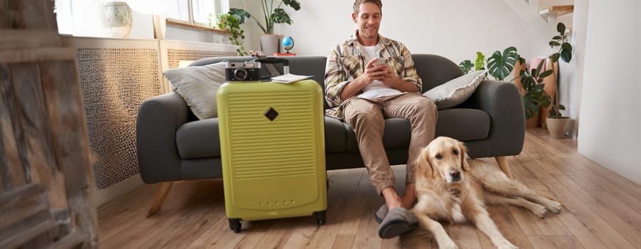 Portrait of guy with a dog, has packed suitcase, camera for going abroad and shooting pictures, dog resting near his legs, man looks at mobile phone and chats online.