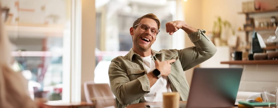 Portrait of handsome, pleased young man shows biceps, flexing muscles, sits in front of laptop in cafe, rejoicing, private coach having a video chat with his client.