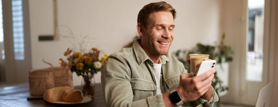 Portrait of smiling charismatic young man, sitting in coffee shop, drinking cappuccino, looking at his mobile phone, reading a message.