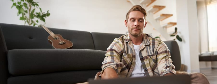 Close up portrait of young man using laptop, looking at computer, sitting on floor with his gadget, browsing websites.