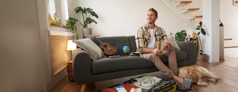 Portrait of man sitting with his dog, packing suitcase for a holiday, going on trip with a pet. Tourism concept