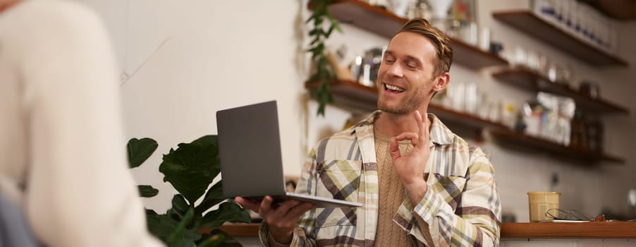 Portrait of young smiling man, coworker joins online meeting from cafe, video chats on laptop, shows okay, ok hand sign in approval, drinking coffee and working remotely.